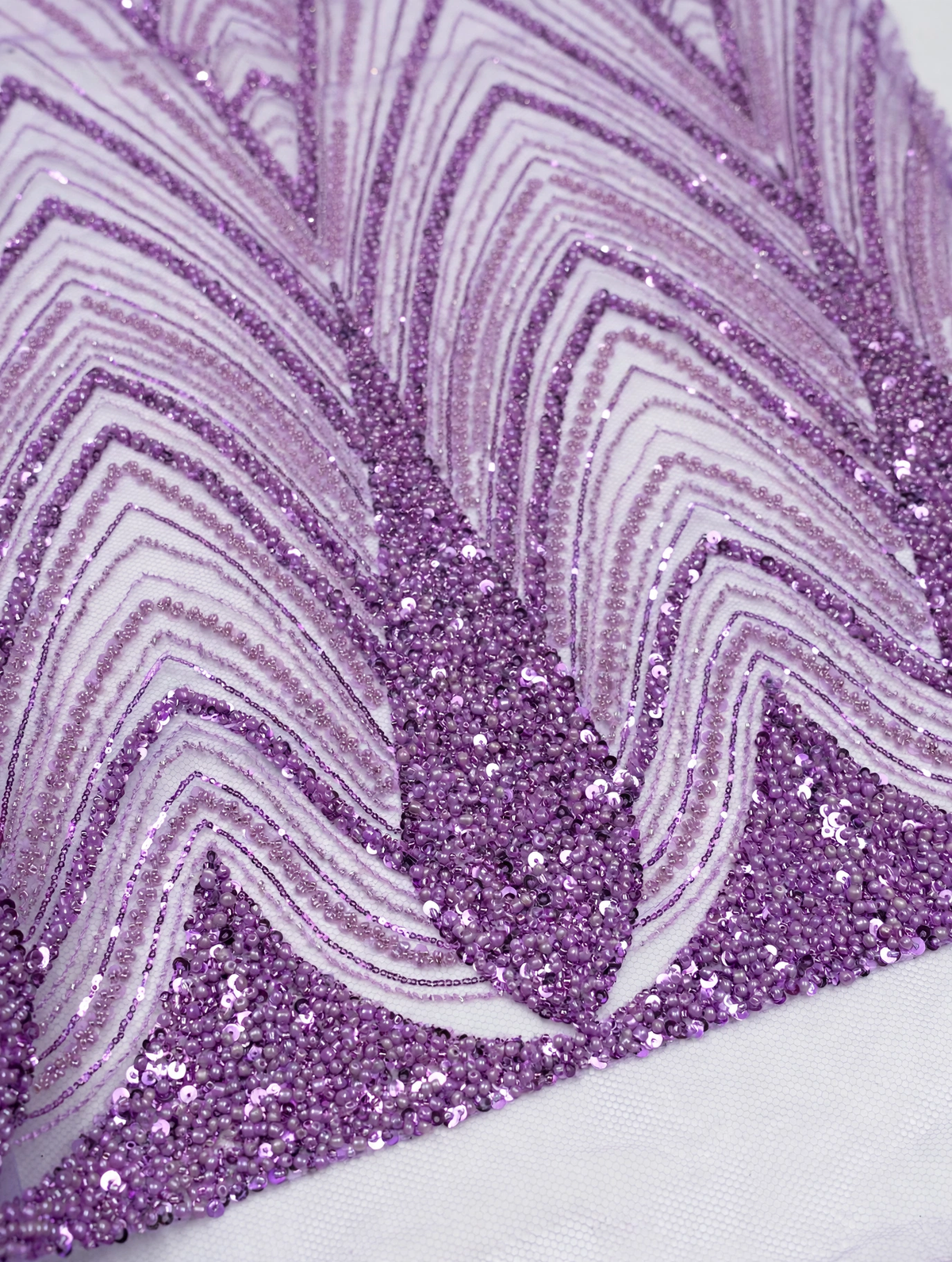 Gros plan des perles et sequins lilas formant les lignes courbes des motifs feuillage sur tulle.